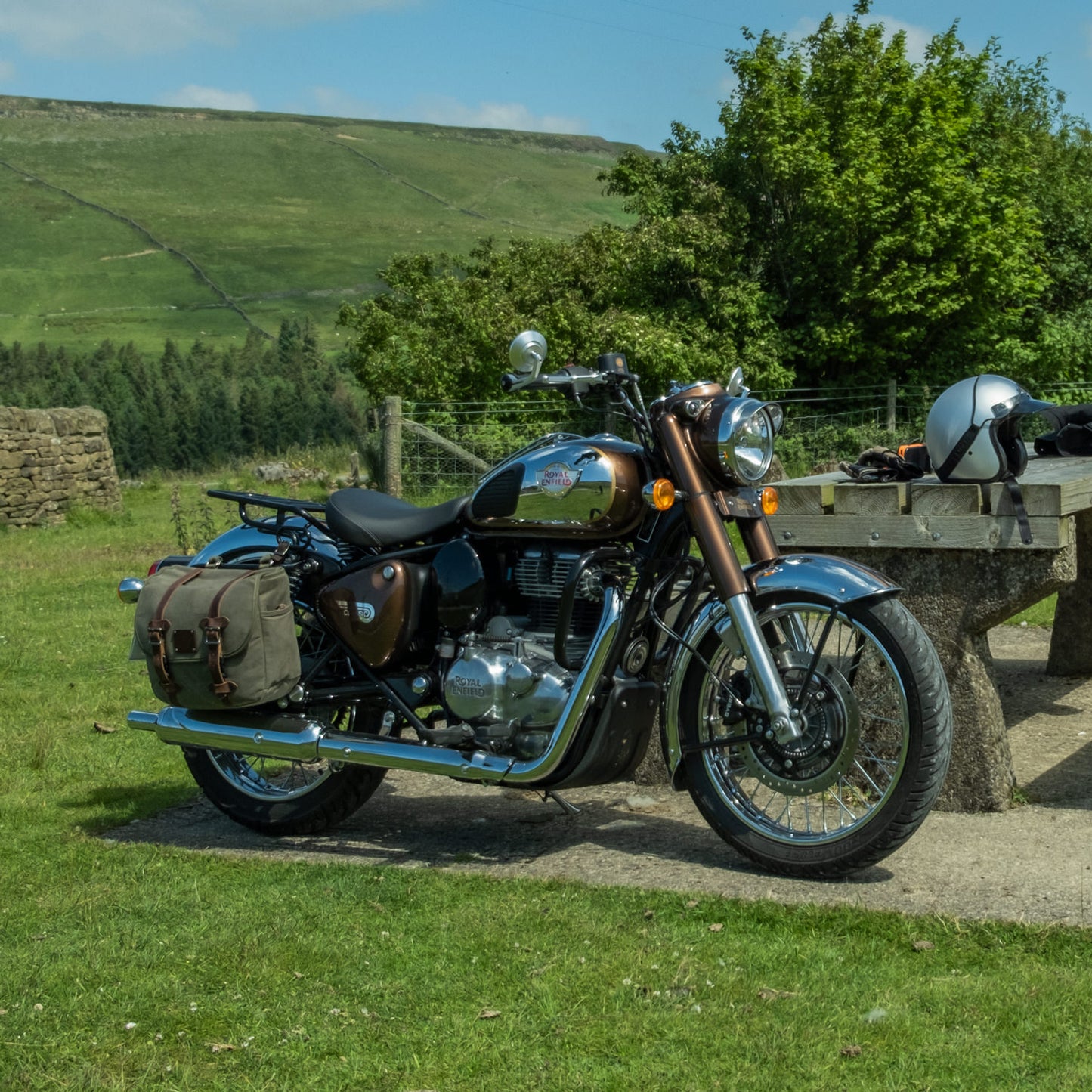 A Royal Enfield Classic 350 Reborn with canvas panniers parked by a bench in the sunlight