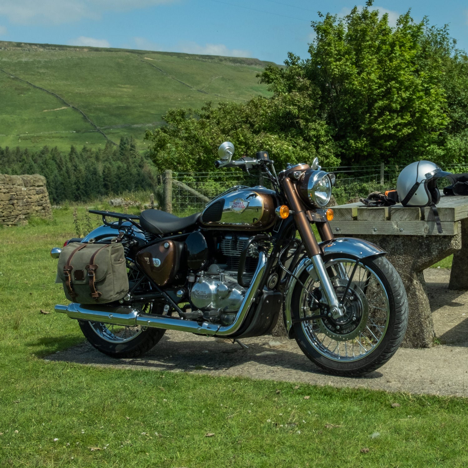 A Royal Enfield Classic 350 Reborn with canvas panniers parked by a bench in the sunlight