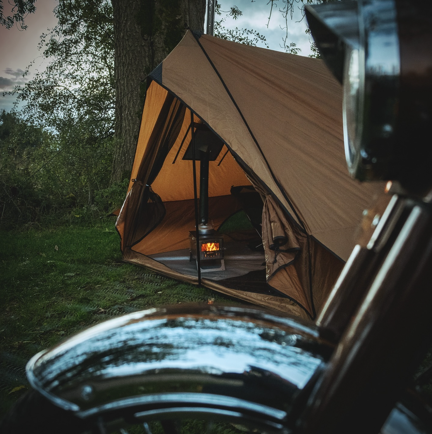 Tent with a stove inside, surrounded by trees and grass, with a classic motorcycle parked in the foreground.