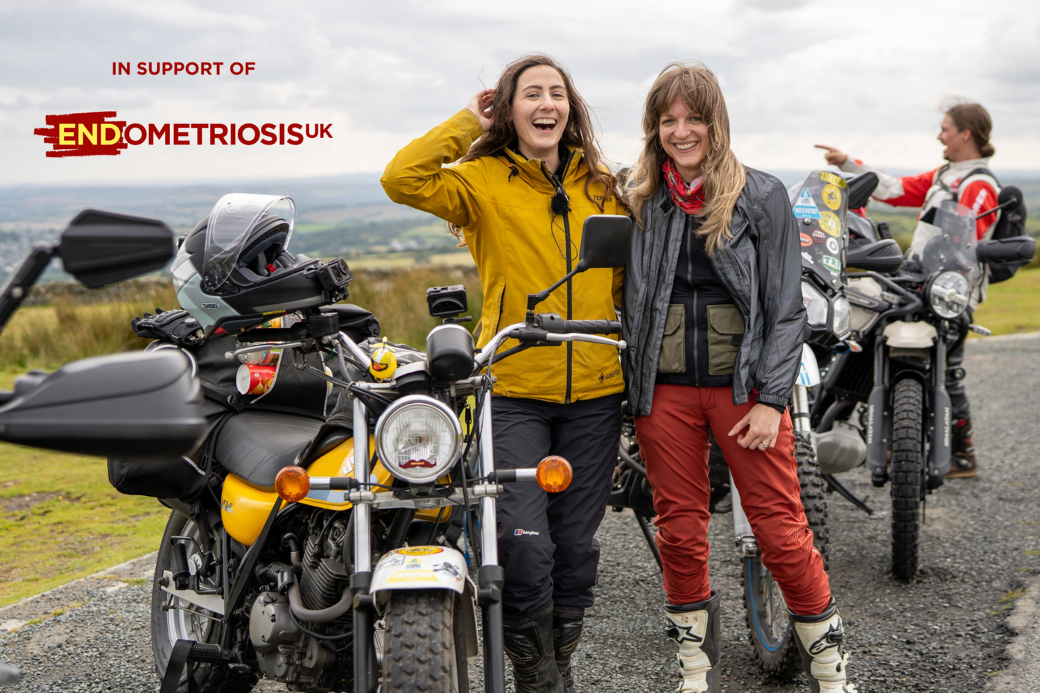Two women standing next to motorcycles with Endometriosis UK support on a scenic road.