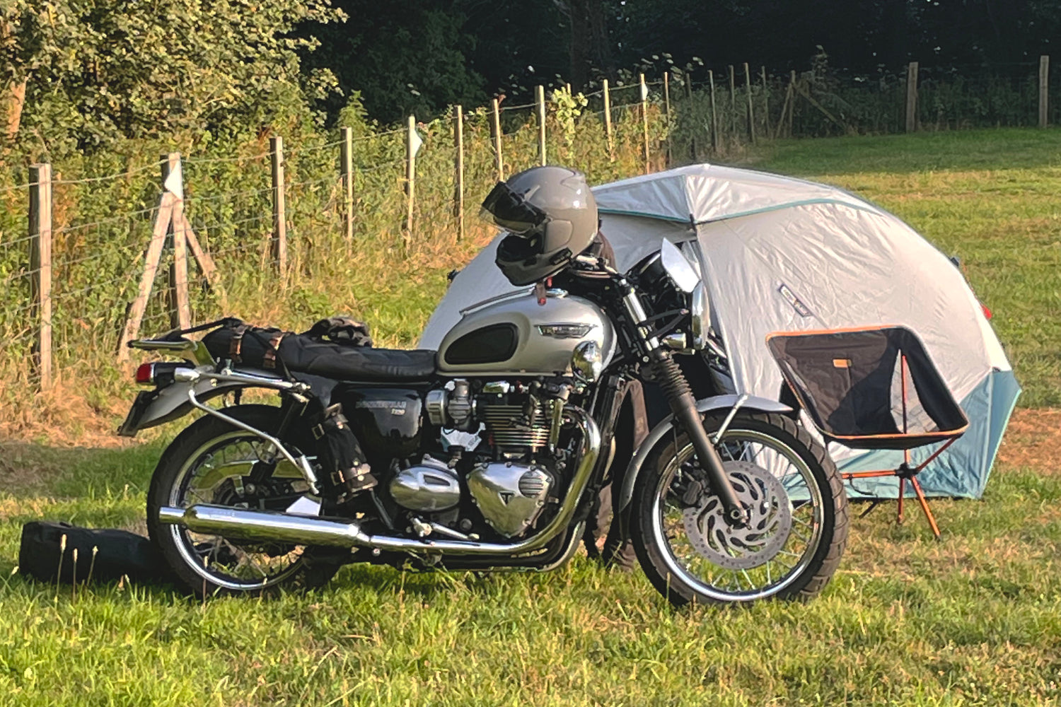 A Triumph Bonneville T120 motorcycle parked next to a tent in a grassy field campsite.